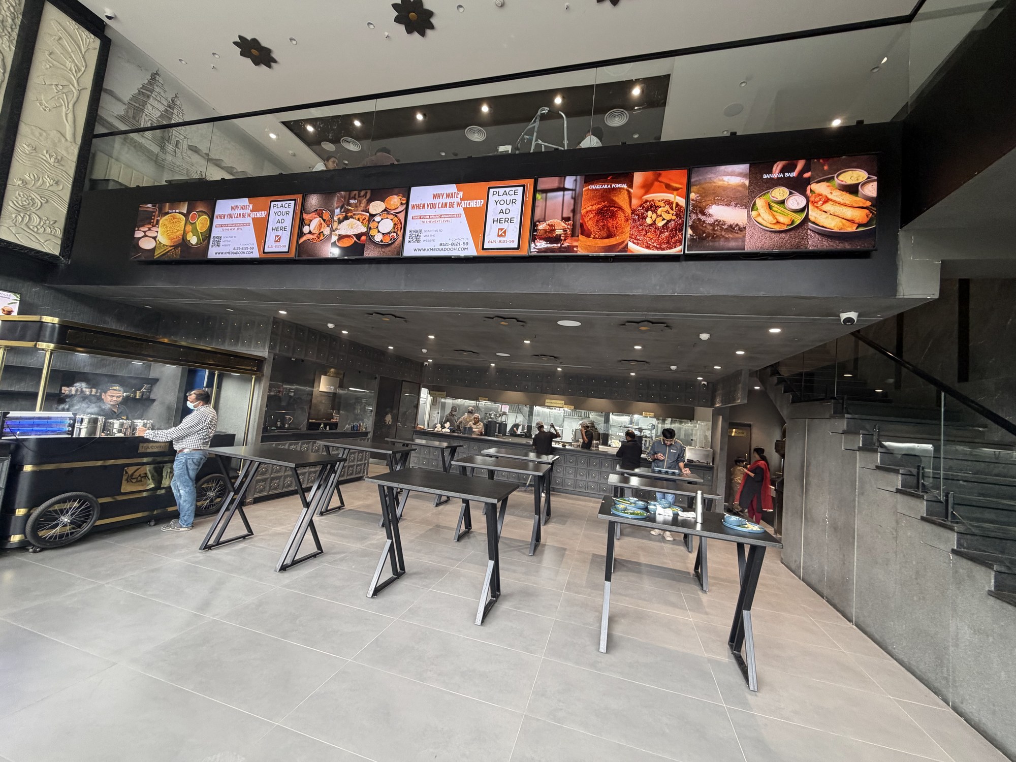 Customers at a restaurant counter beside a KMEDIA digital standee displaying ads
