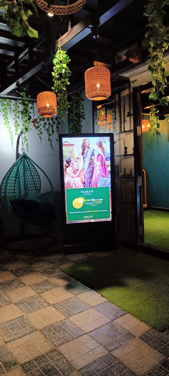 Vaarahi Silks ad on KMEDIA standee at a garden-style cafe